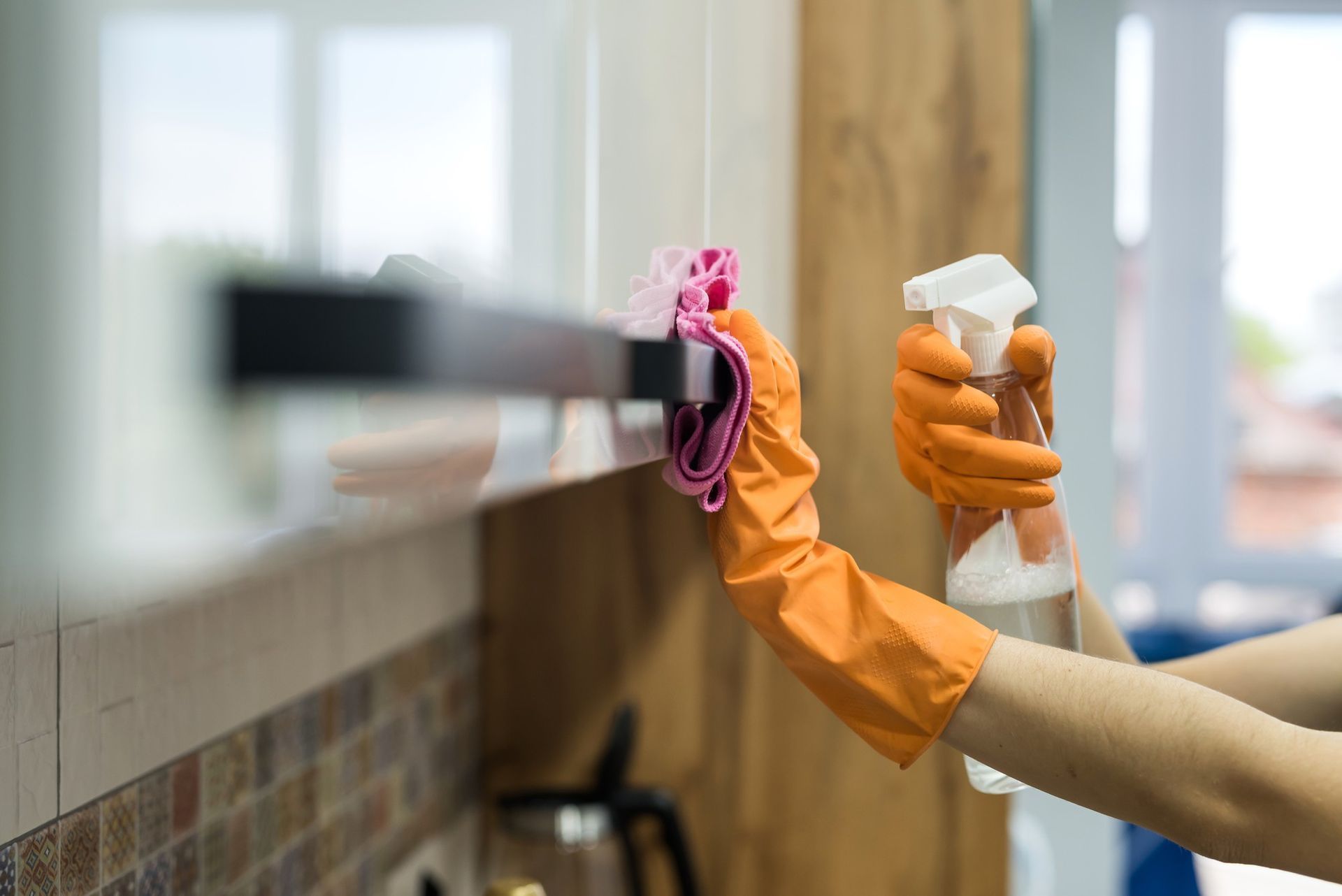 Person wearing orange gloves sprays and wipes a kitchen cabinet with a pink cloth.