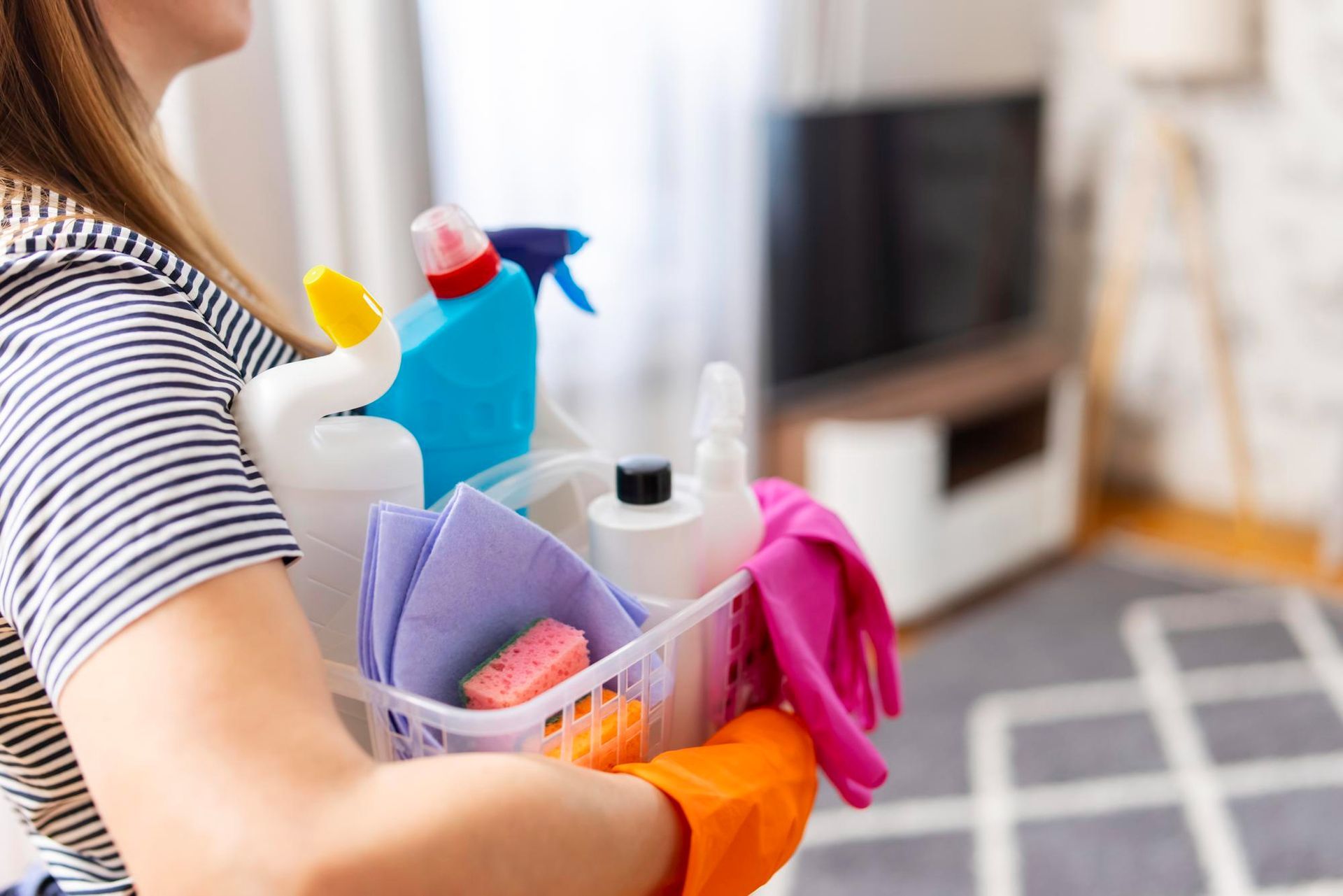 Woman holding a basket filled with cleaning supplies, including bottles, rags, and gloves. Living room in background.