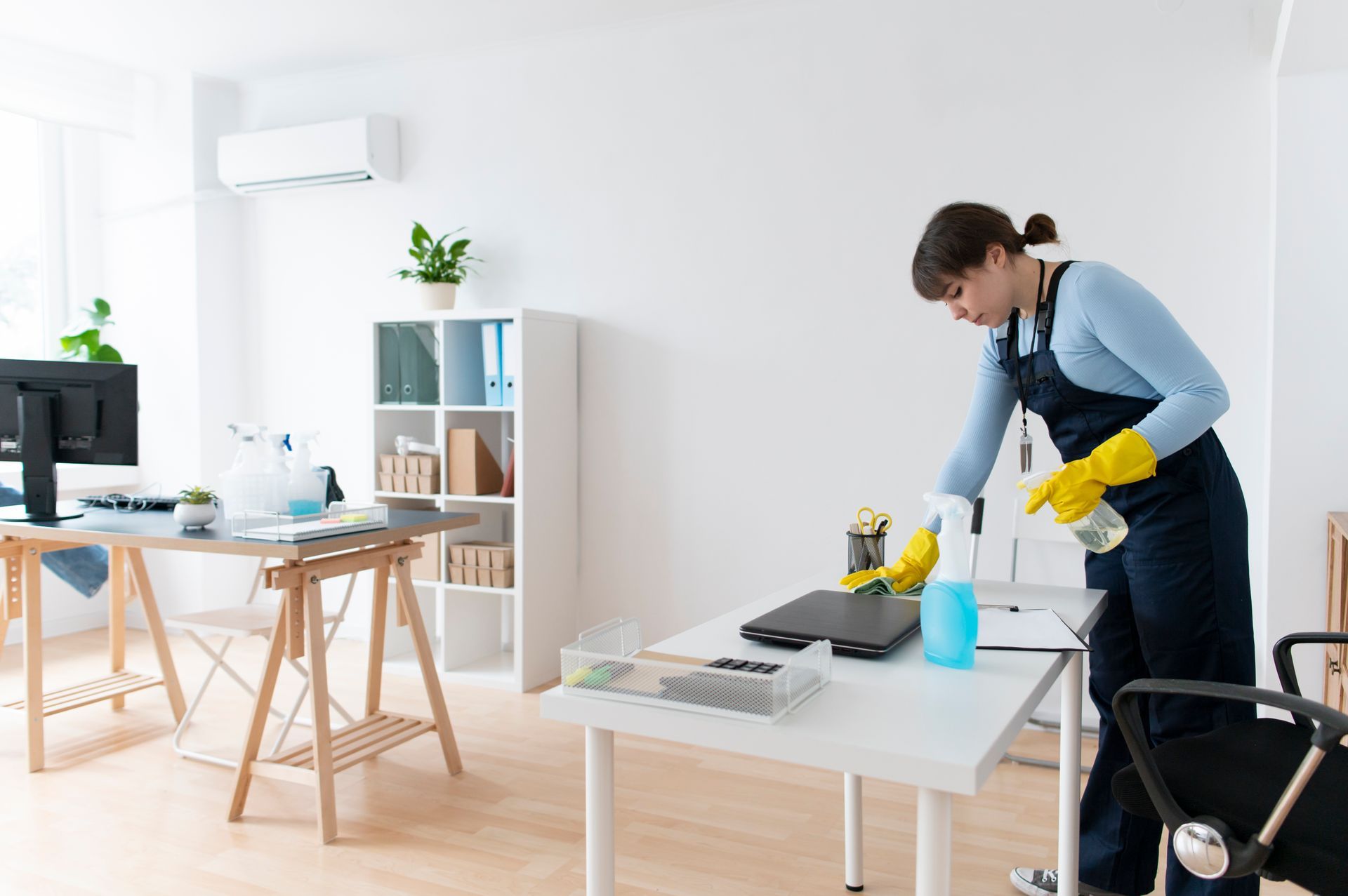 Person wearing gloves cleaning a desk in a bright office with a computer and bookshelf.