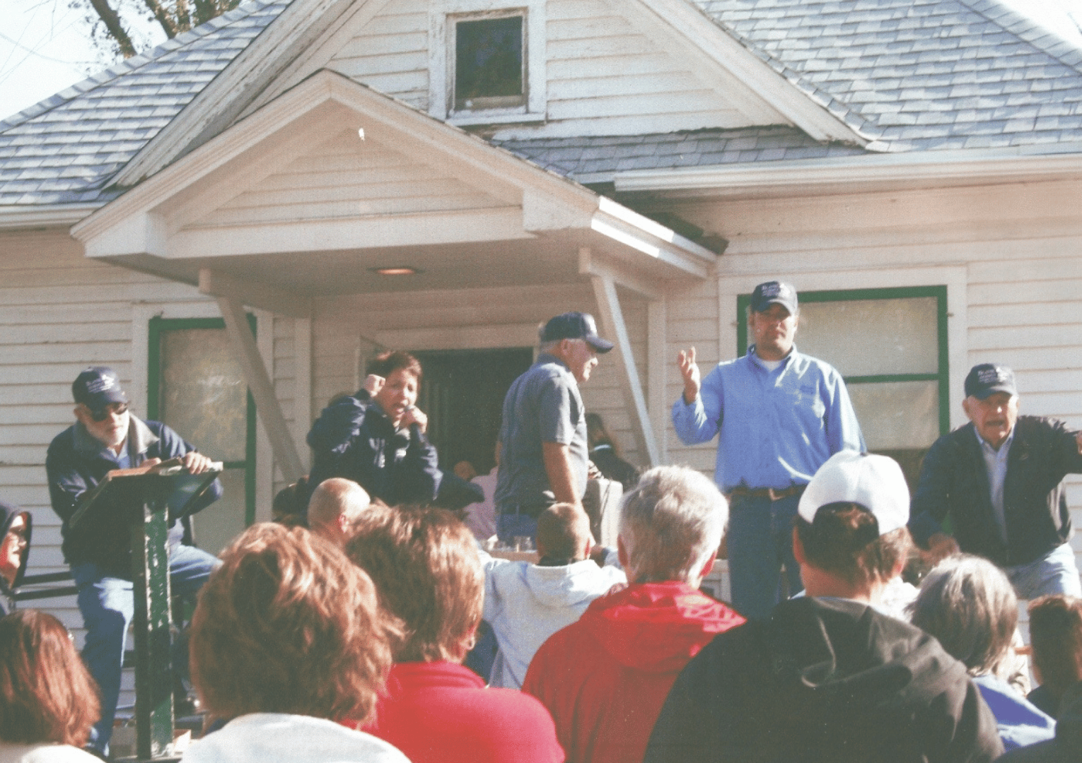 People at a house auction; a man gestures with two fingers, others watch, light-colored house in background.