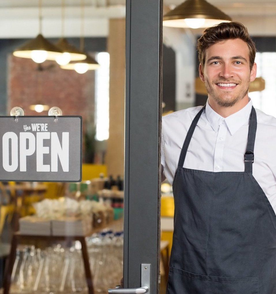 Man in apron smiles, standing in doorway of open restaurant with “We’re Open” sign.