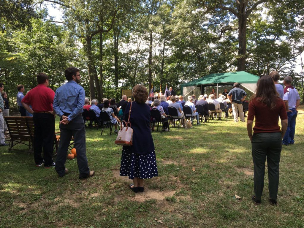 People gathered outdoors in a park for an event, with chairs and green umbrellas under trees.