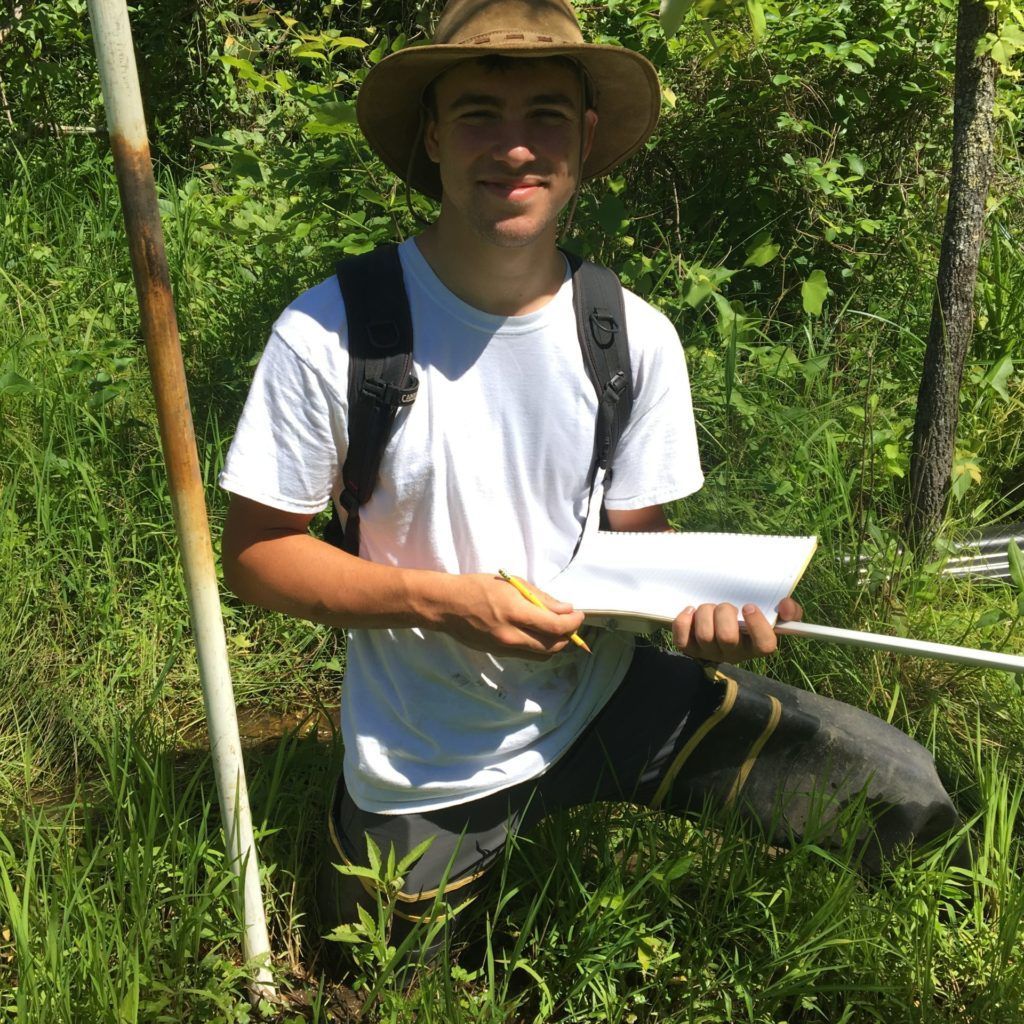 Man kneeling in tall grass outdoors, wearing a hat and backpack, holding a white notebook and pen.
