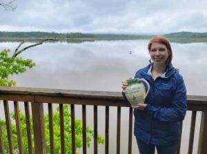 Person in a blue jacket holding a jar on a wooden deck overlooking a calm lake.