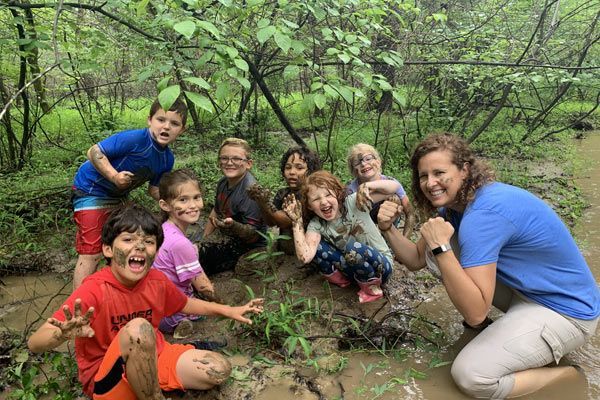 Group of children posing in a muddy forest stream, smiling and crouching among green trees.