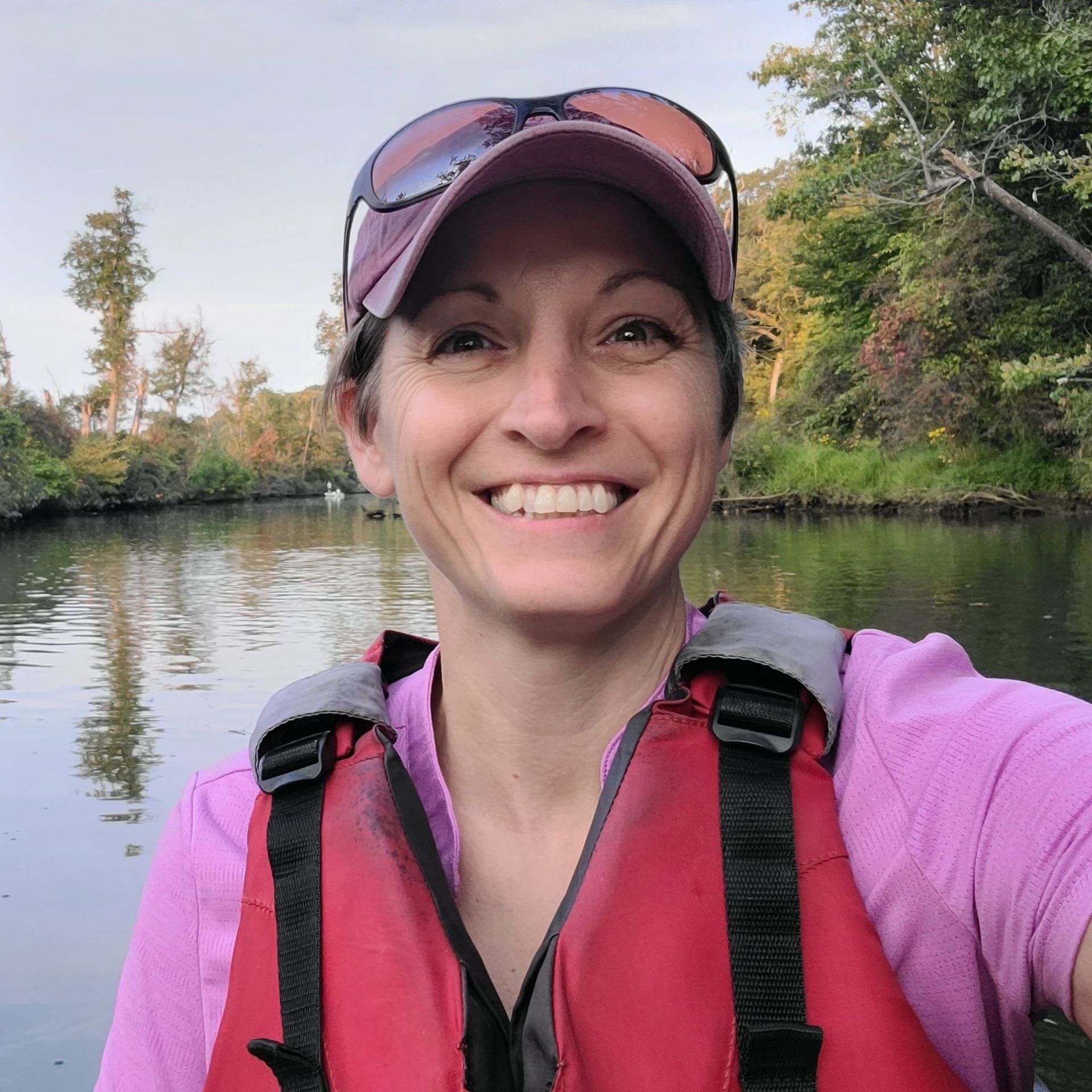 Smiling woman in a pink life vest and cap on a calm river with trees in the background