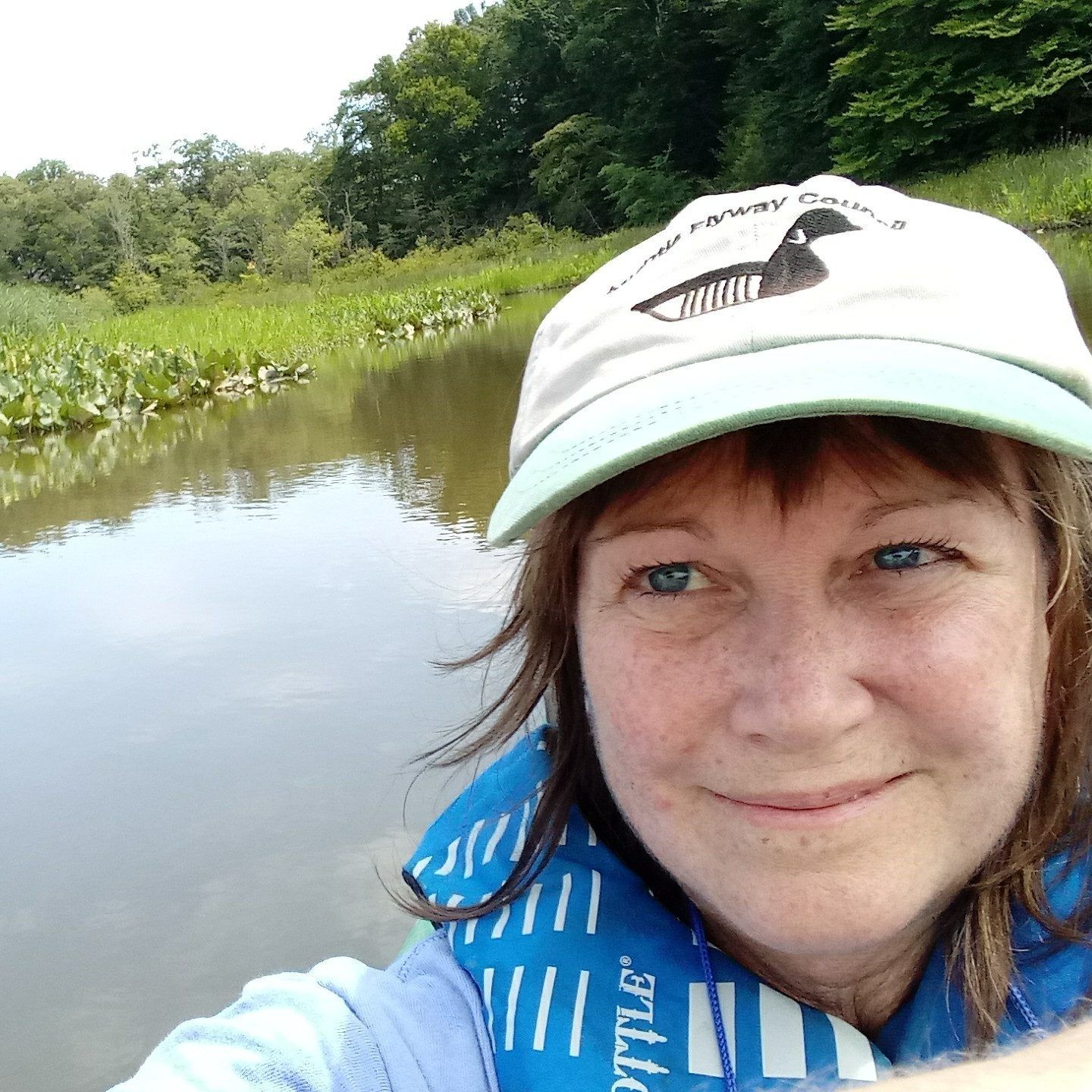 Person taking a selfie by a calm river with green trees and grassy banks in the background