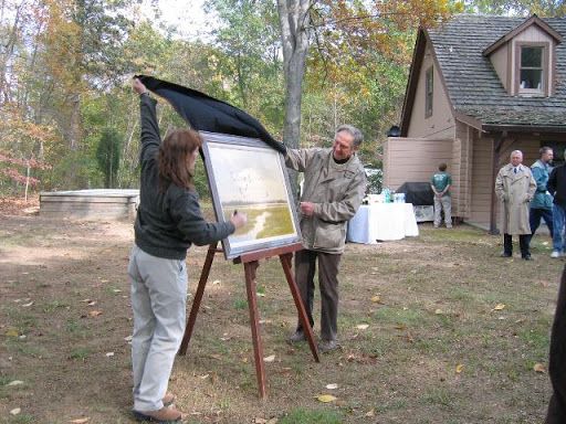 Two people at an easel outdoors  unveiling a painting, near a cabin, with others standing nearby in a wooded yard.