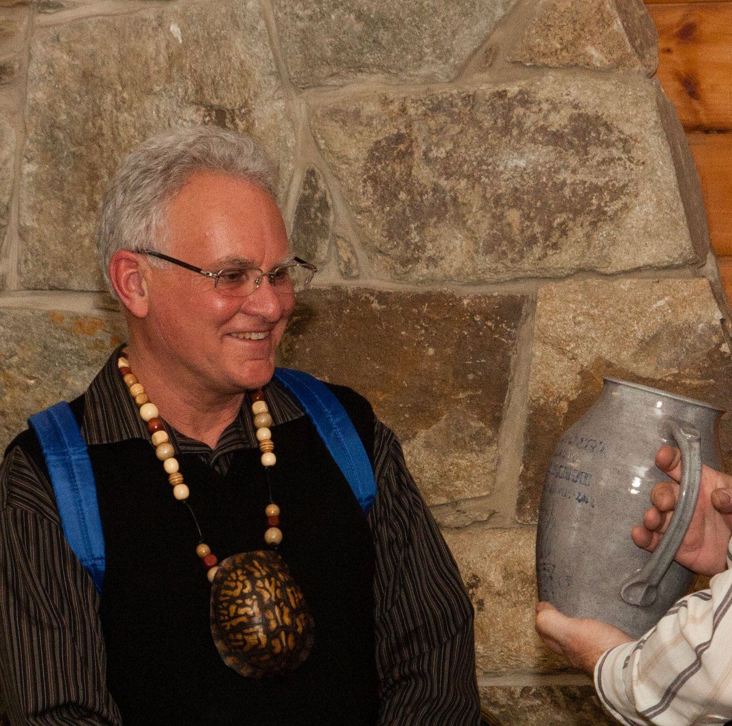 Man smiling at a stone wall, wearing glasses and a beaded necklace, receiving a jug