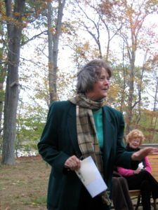Woman in a green jacket and scarf holding papers outdoors, with another person seated behind her.