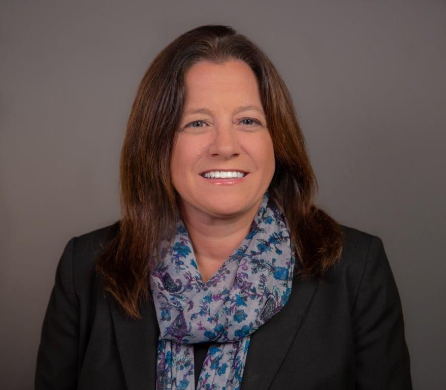 Smiling woman in a black blazer and blue patterned scarf against a gray background.