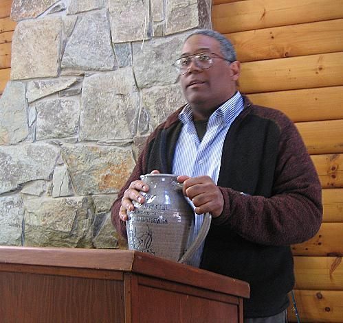 Man speaking at a podium, holding a decorative jug in front of a stone and log wall