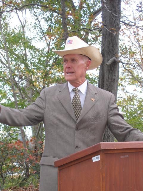 Man in a cowboy hat and suit speaking at a podium outdoors among trees