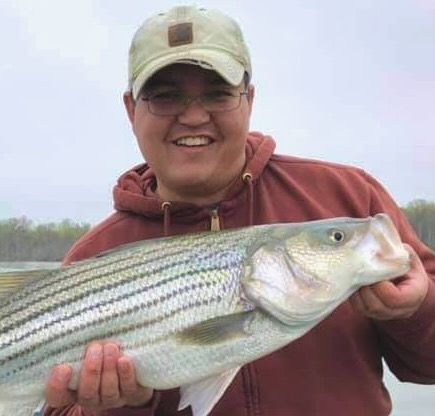 Person smiling and holding a large striped fish outdoors by the water