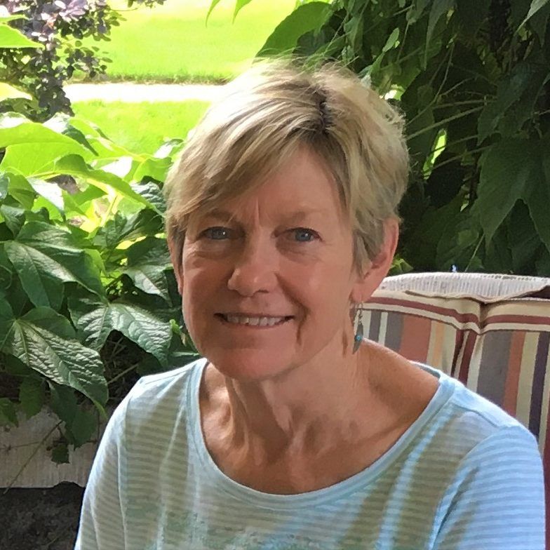 Smiling blonde woman seated on a porch with lush green plants in the background