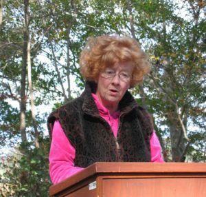 Woman speaking at a podium outdoors, wearing a bright pink shirt and dark vest, with trees in the background