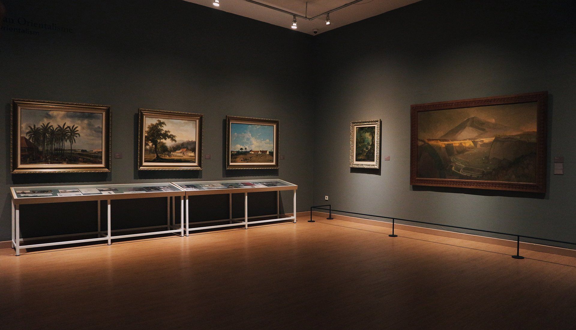 Dimly lit art gallery with landscape paintings displayed on dark walls and a long glass display case.
