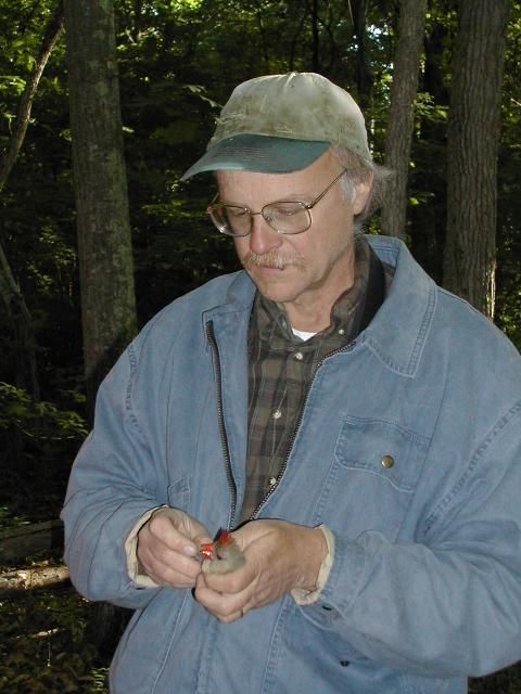Man in a blue jacket and cap holding a bird outdoors among trees.