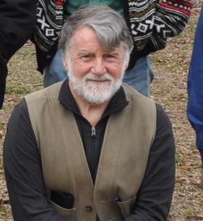 Smiling man with gray hair and beard in a tan vest, seated outdoors with people behind him