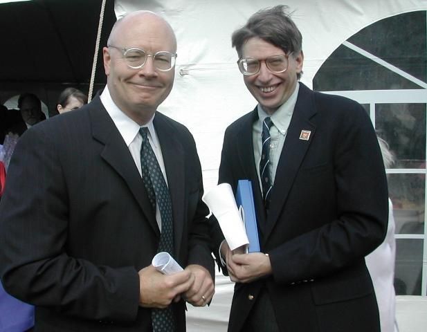 Two men in suits smiling and holding papers at an outdoor event tent