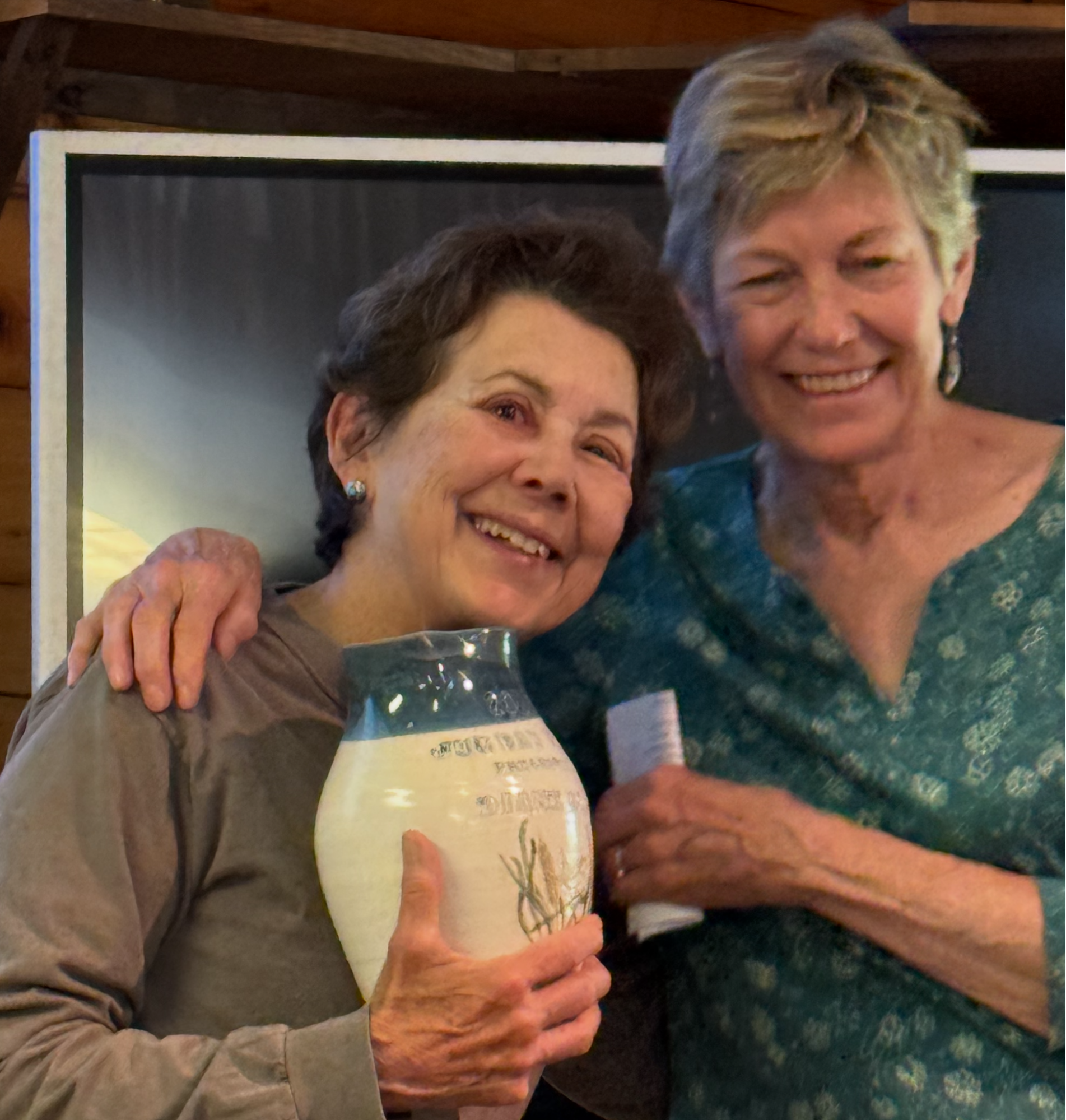 Two smiling women posing indoors, one holding a decorative ceramic jar