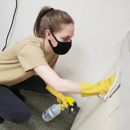 Person in a mask and yellow gloves cleaning a wall with spray bottle and cloth