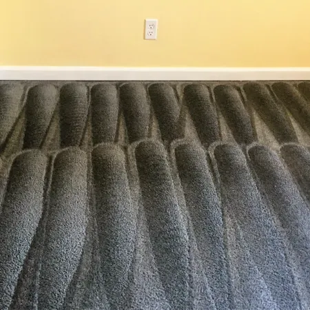 Gray plush carpet with wavy ridges against a yellow wall and white baseboard, with an electrical outlet above.