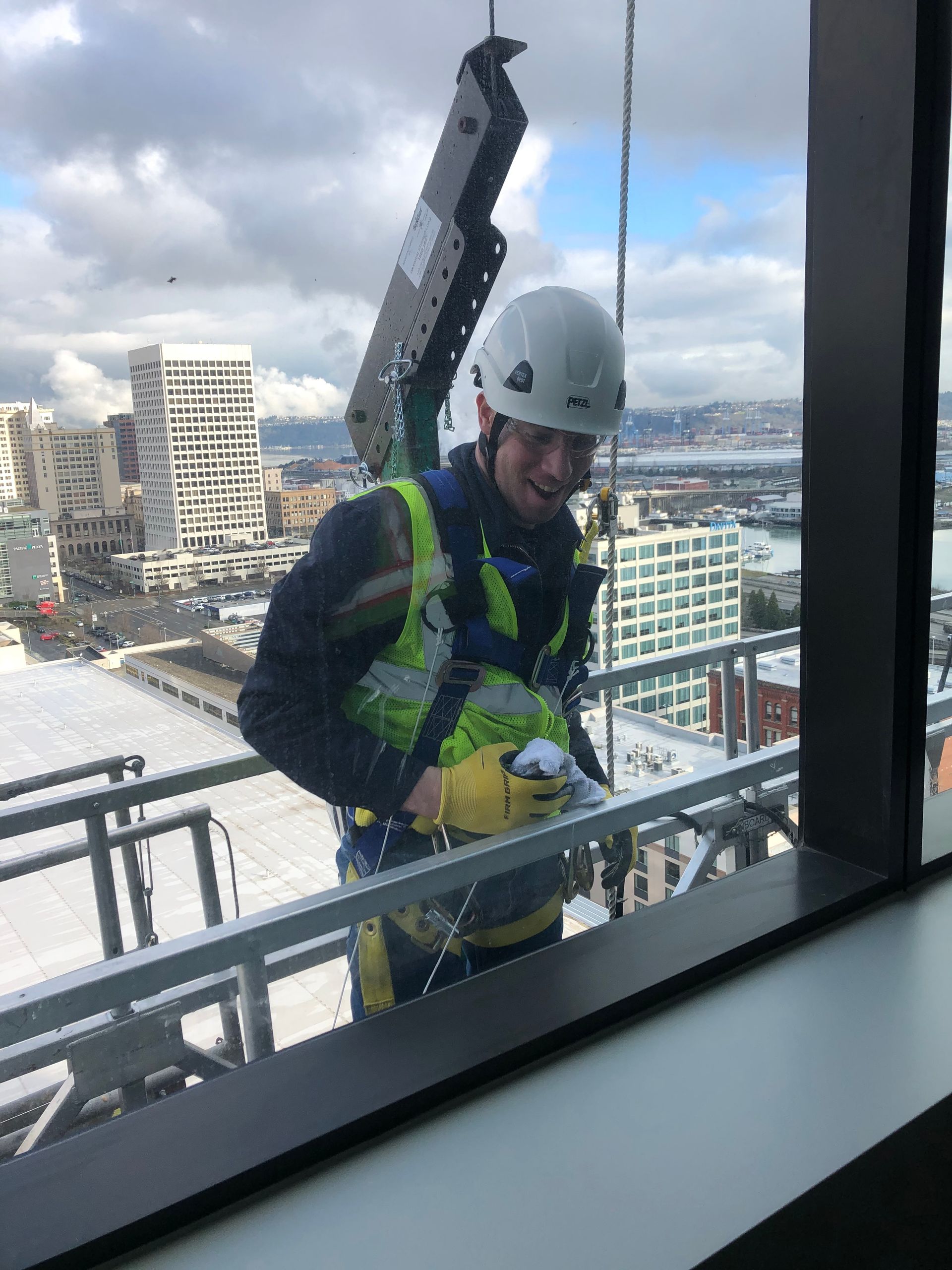 Construction worker in hard hat and safety harness on a high-rise balcony, with city and water behind.