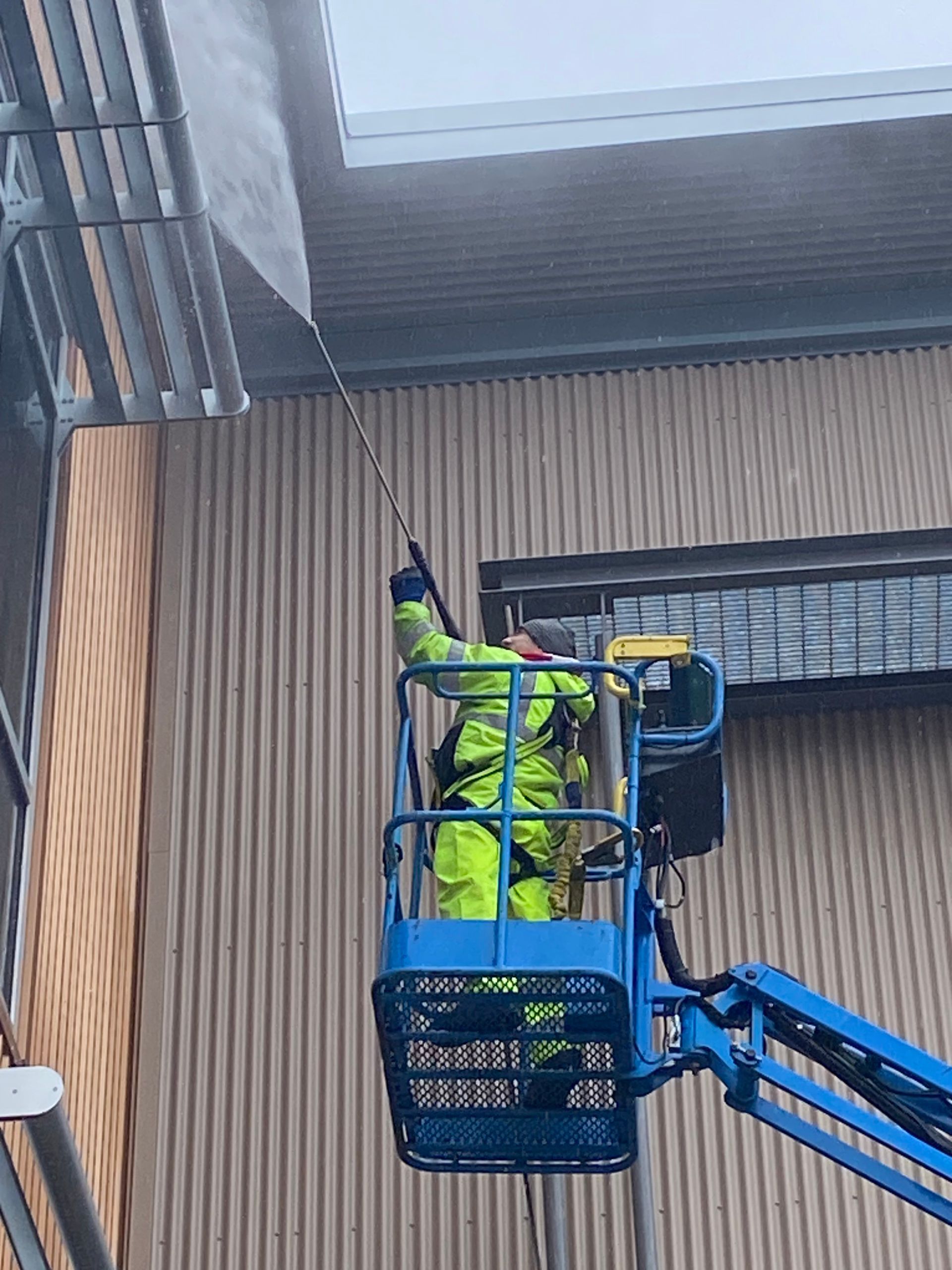 Worker in neon safety gear in a blue lift, reaching up near a building wall and roofline.