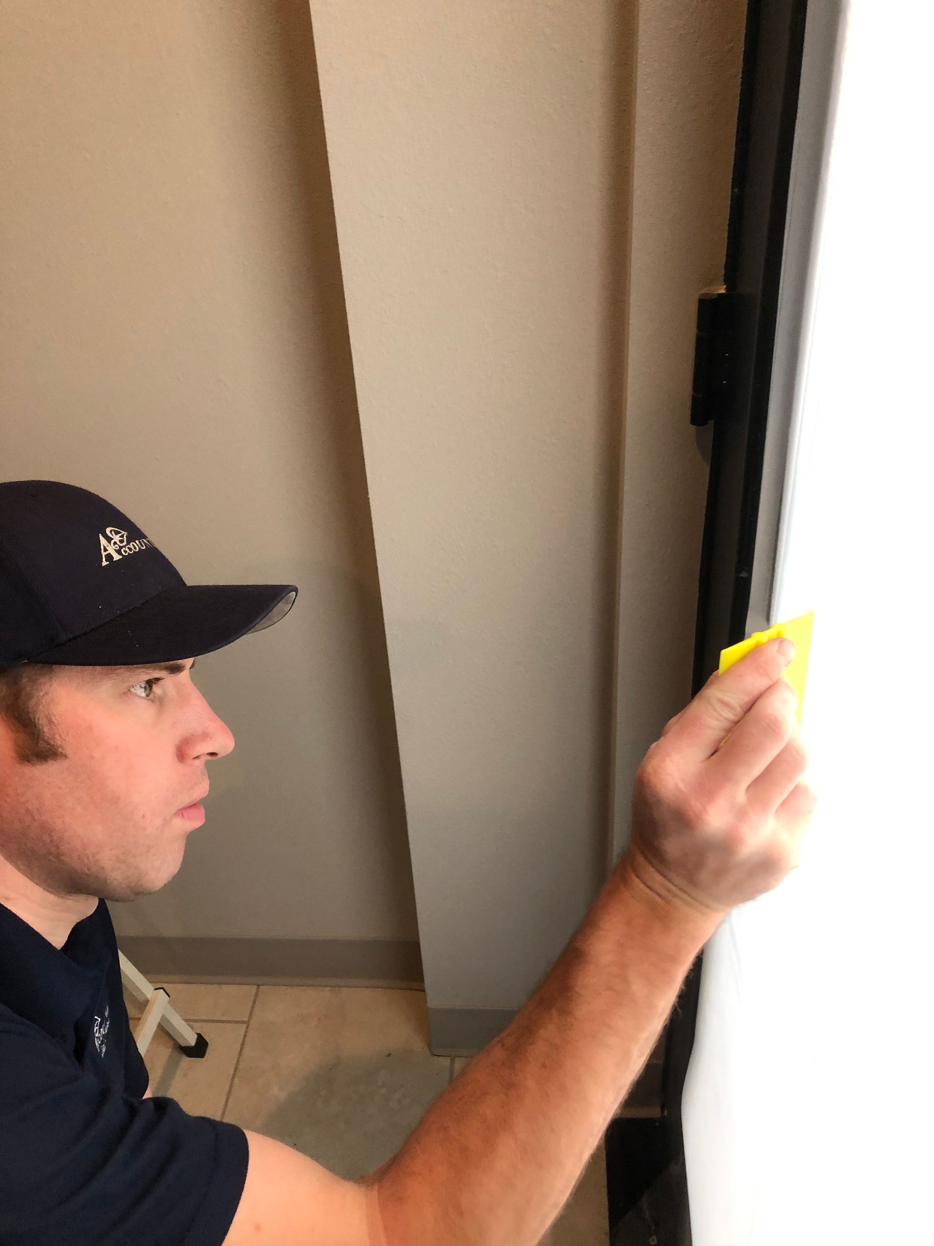 Man in a dark cap using a yellow scraper on a wall near a window
