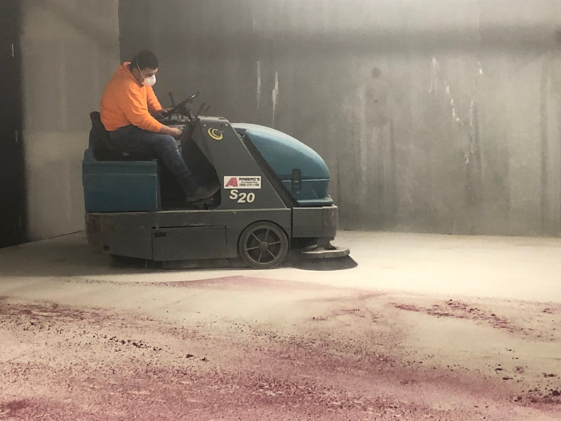 Worker in orange shirt operating a blue floor scrubber in a concrete warehouse.