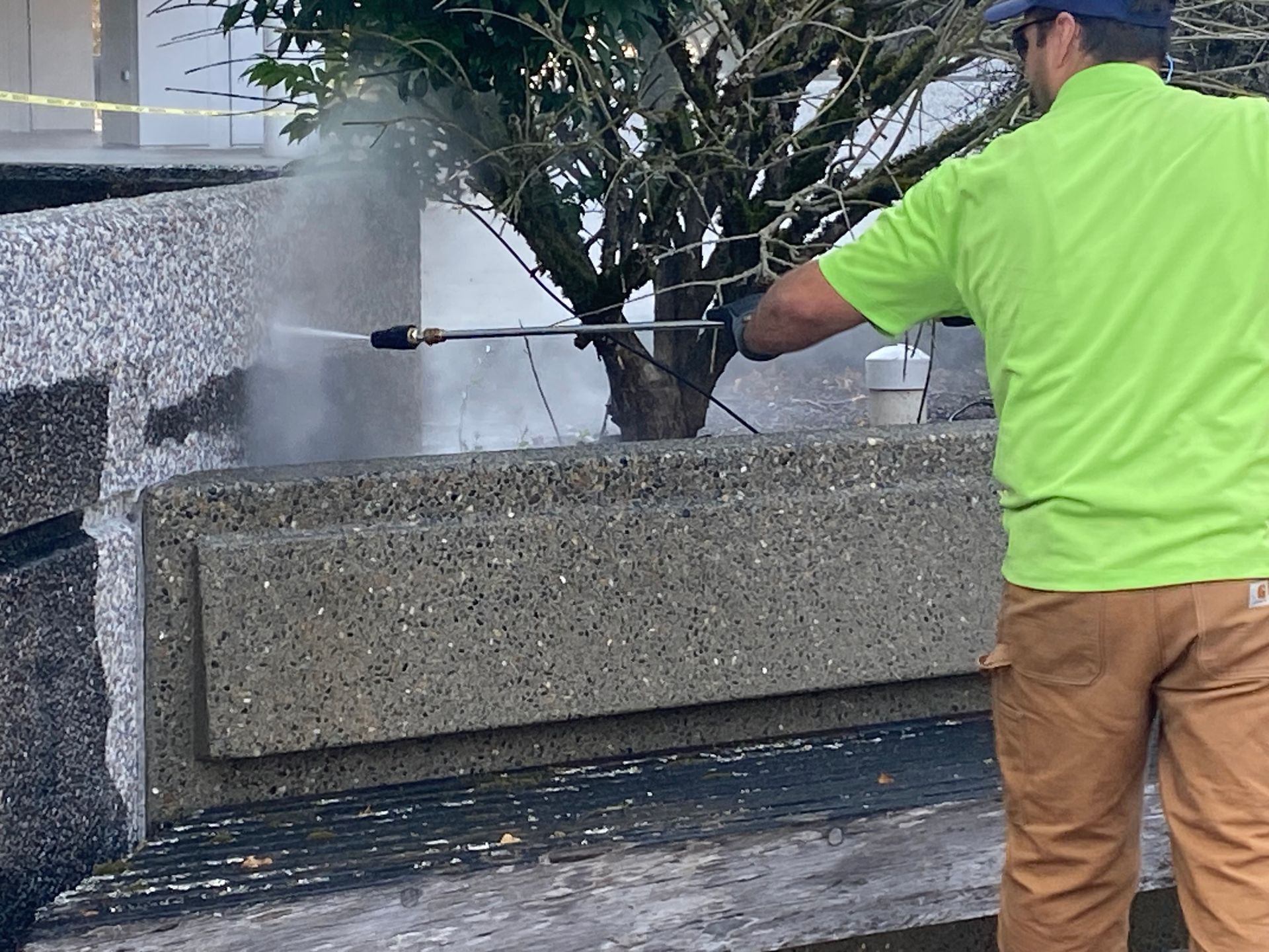 Worker in neon green shirt pressure-washing a stone planter beside a tree.