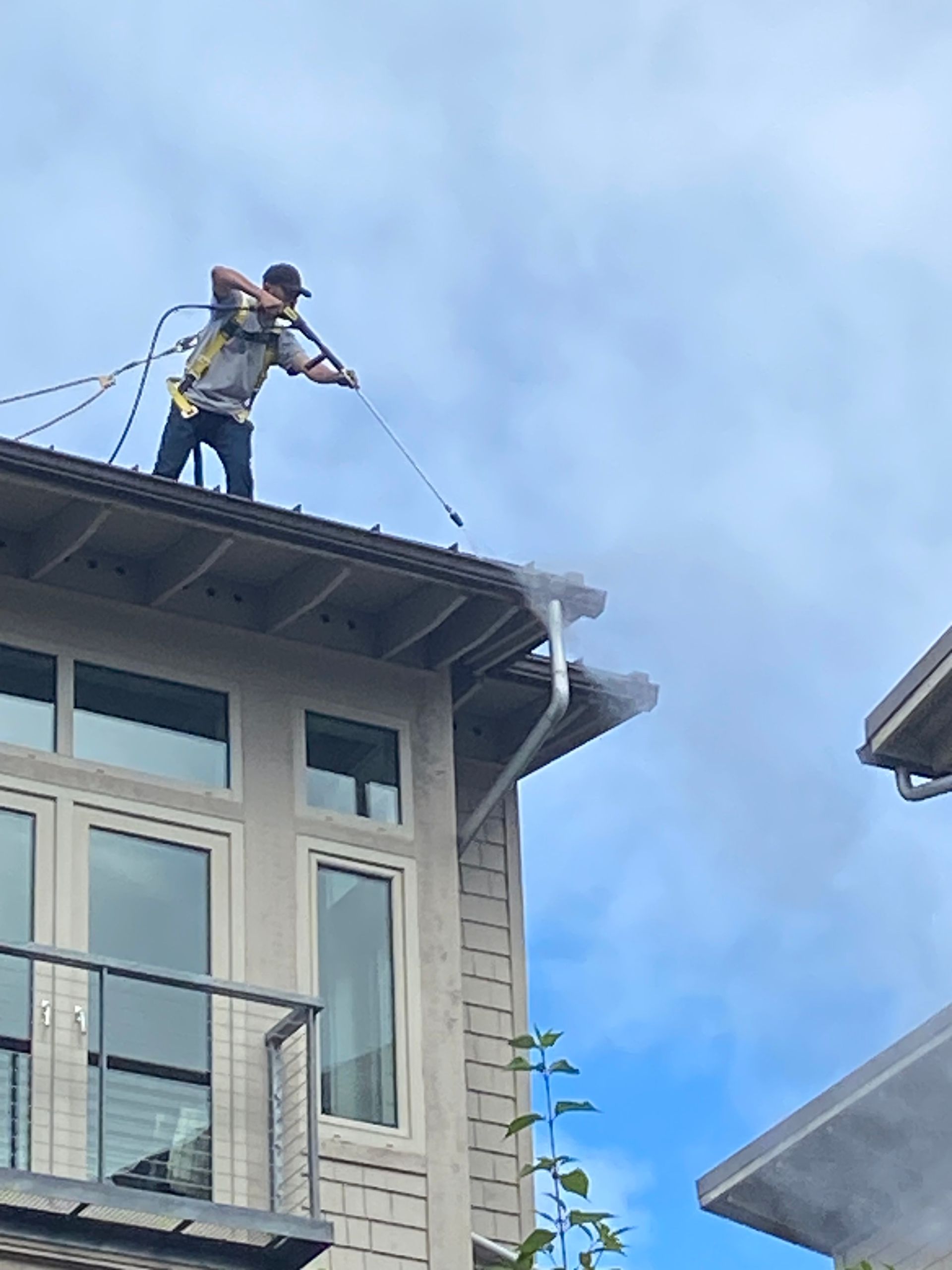 Worker on a rooftop power washing the edge of a house under a cloudy sky
