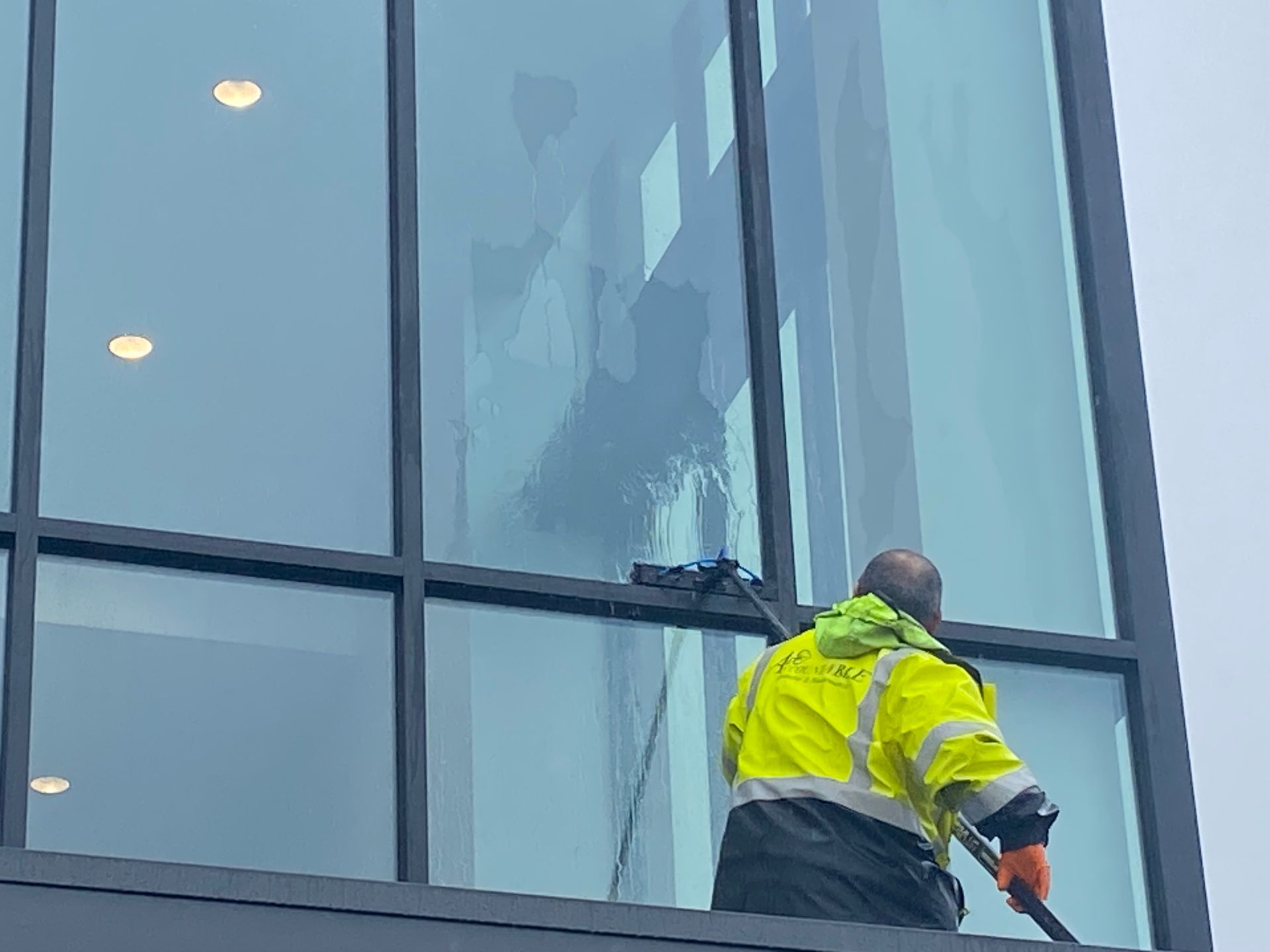 Worker in neon yellow jacket cleaning a tall glass building exterior with a pole.