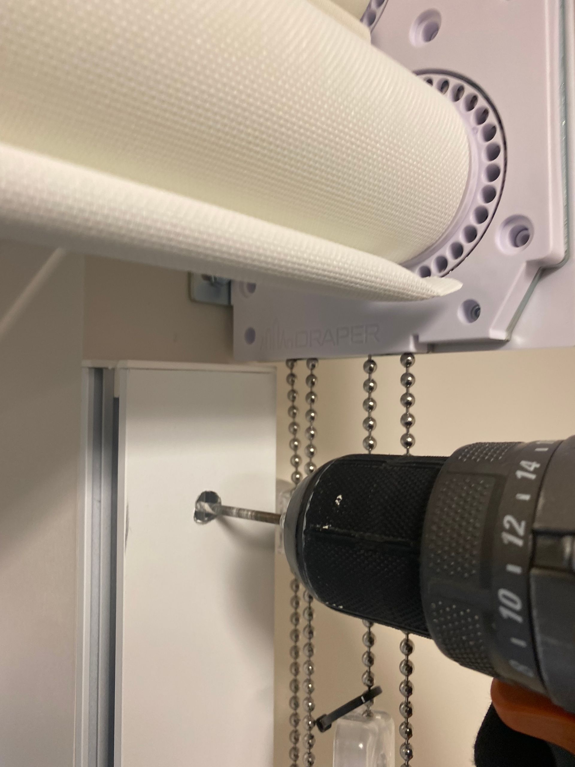 Blind cord attached to a wall-mounted bracket beside a black drill, under a window blind roll-up housing