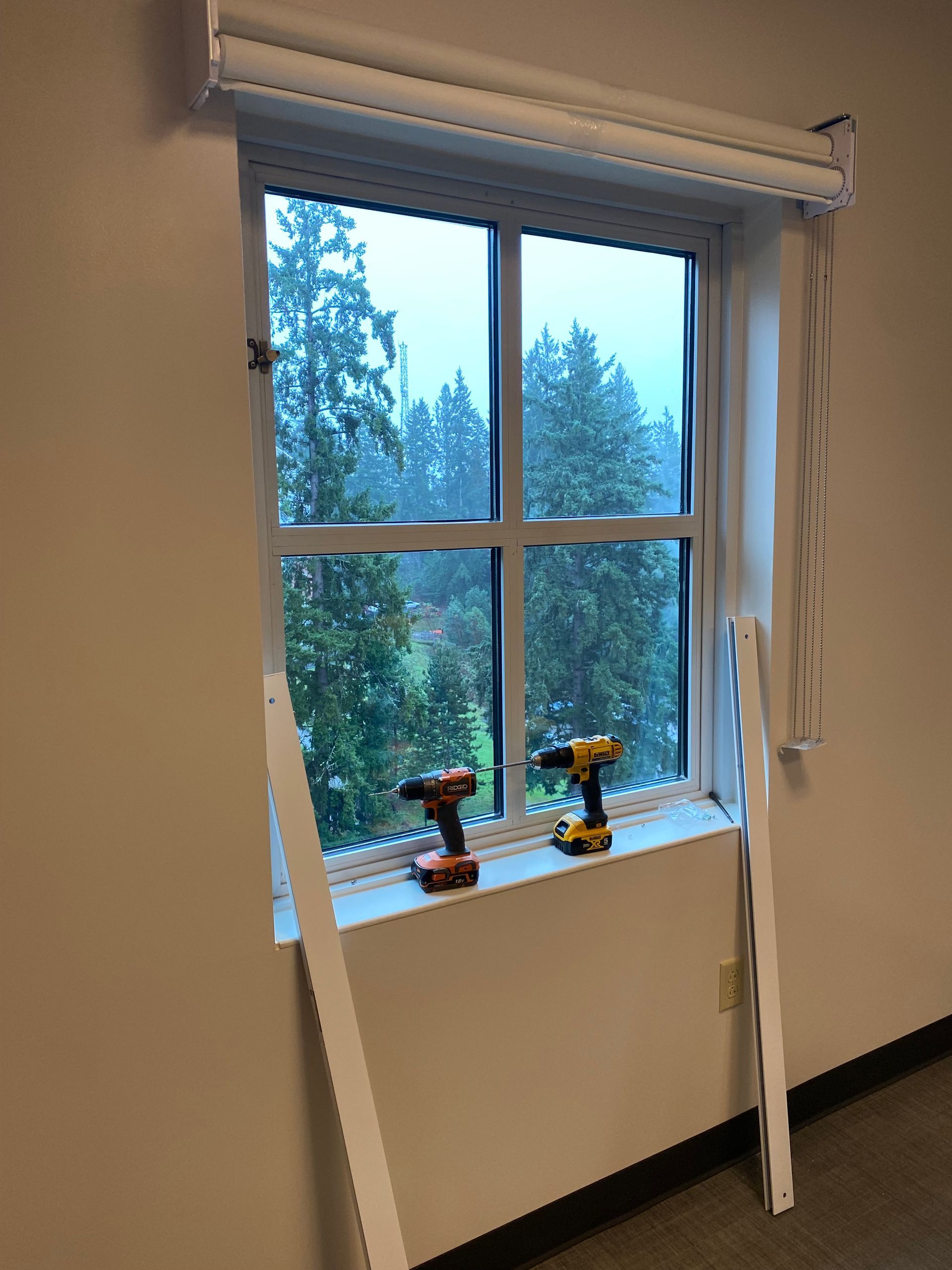 Open window with a white blind, two small drills on the sill, and trees outside.