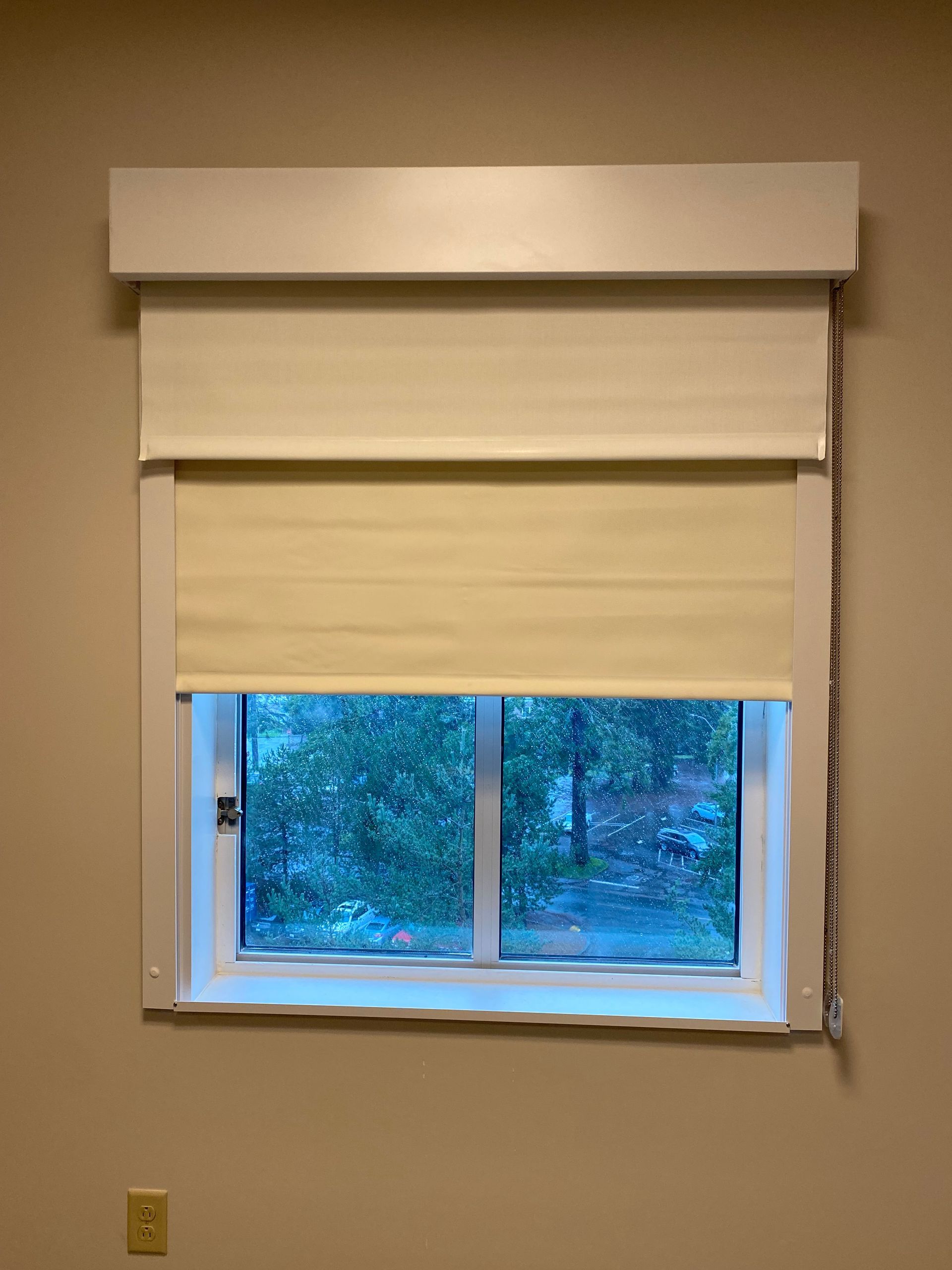 Window with beige blinds and valance, showing a rainy blue-green outdoor view.