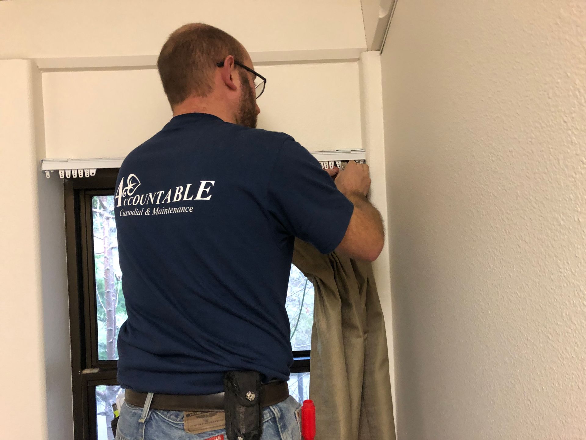 Worker hanging beige curtains beside a window in a room, wearing a navy shirt