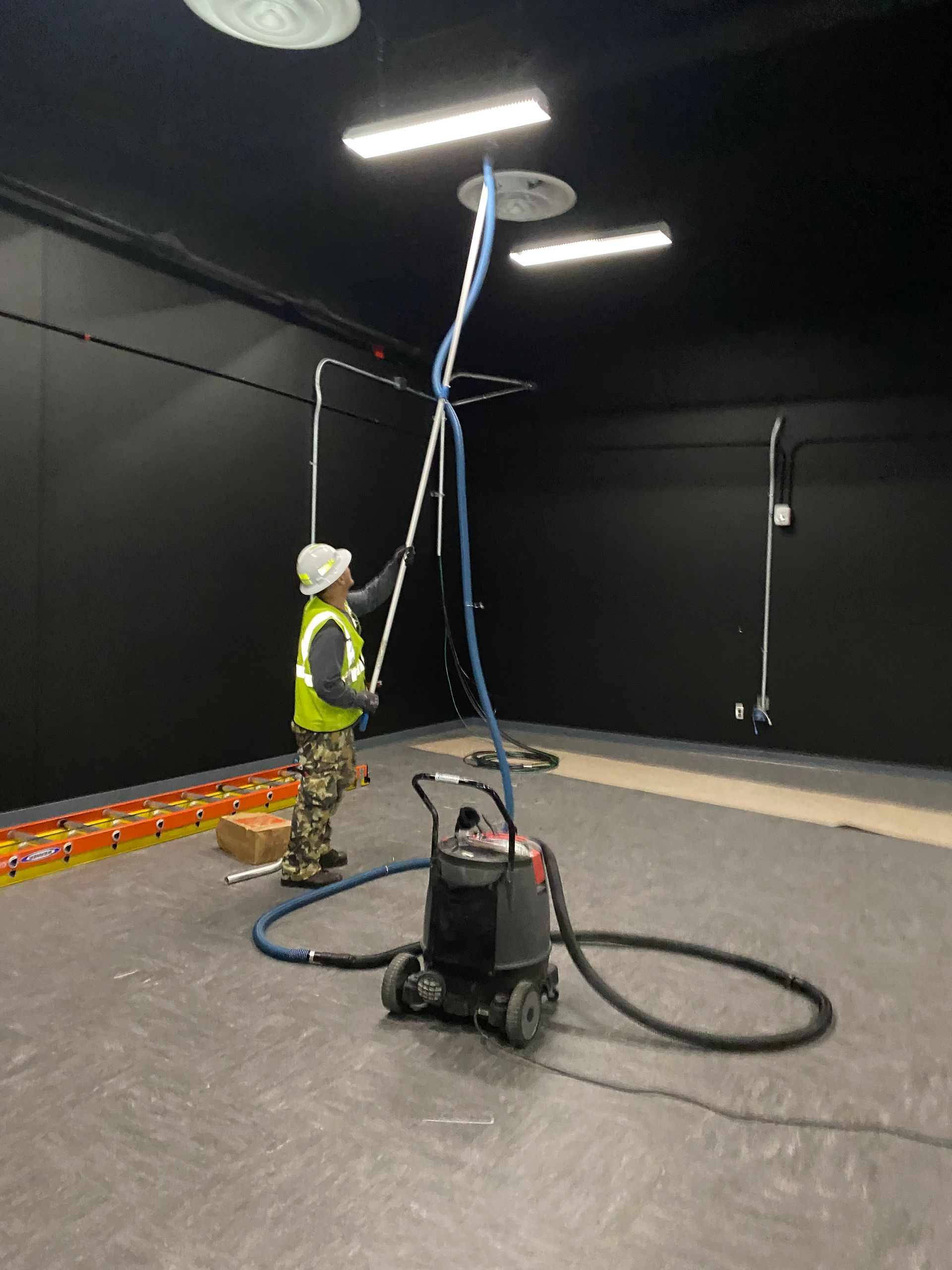 Worker in hard hat and safety vest handling a tall blue cable in a dark indoor room with equipment.