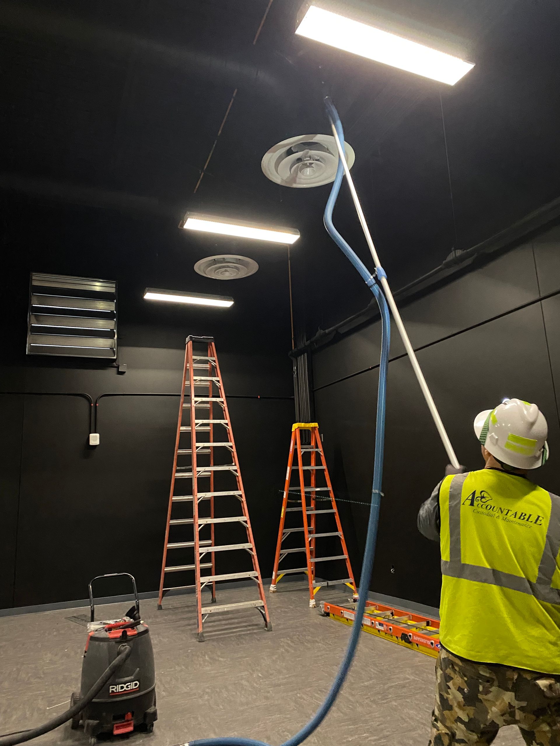 Worker in a yellow vest sprays the black ceiling while standing by ladders in a dark room.