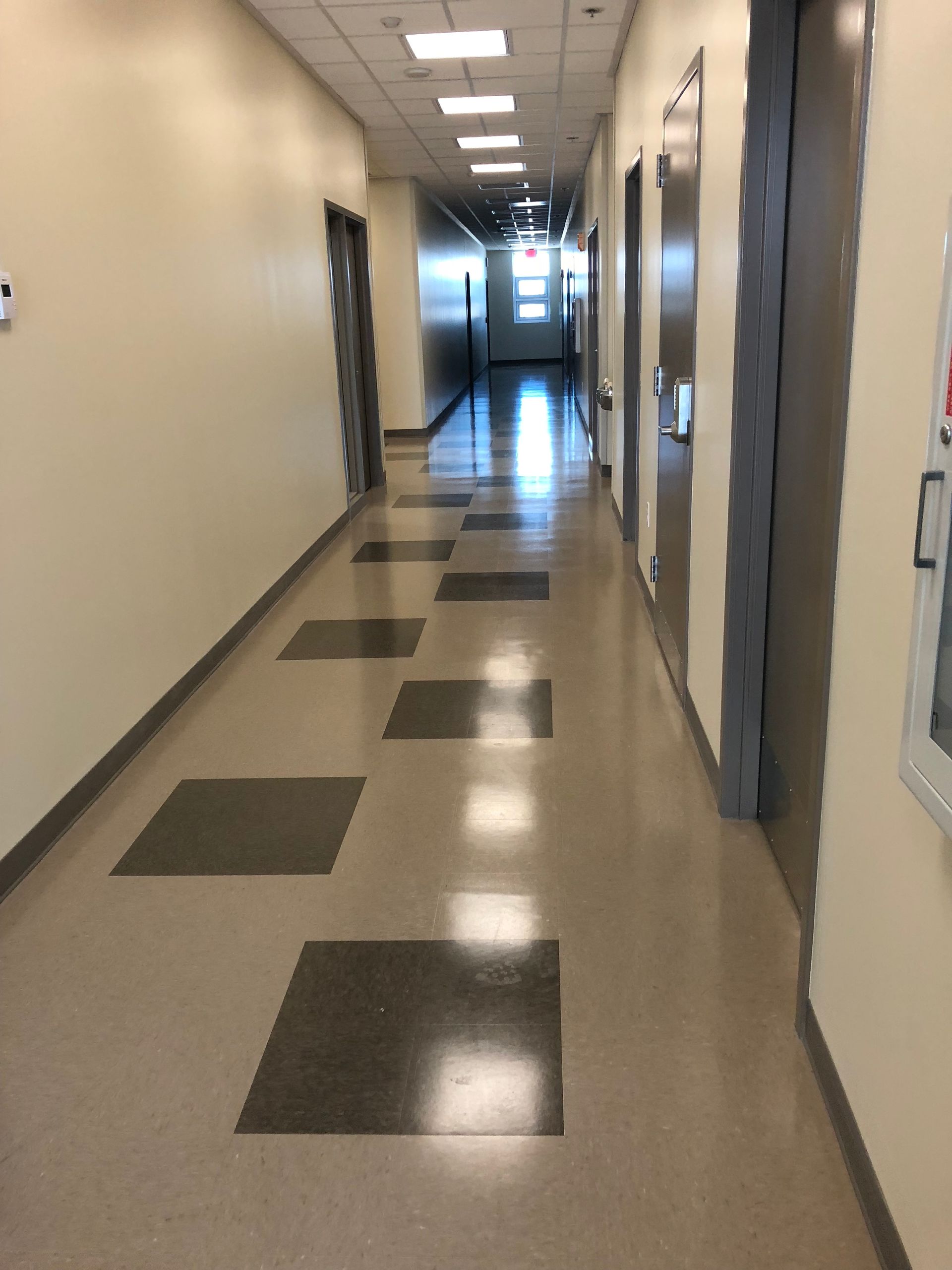Empty hallway with beige walls, gray floor tiles, and bright overhead lights leading to a door at the end.