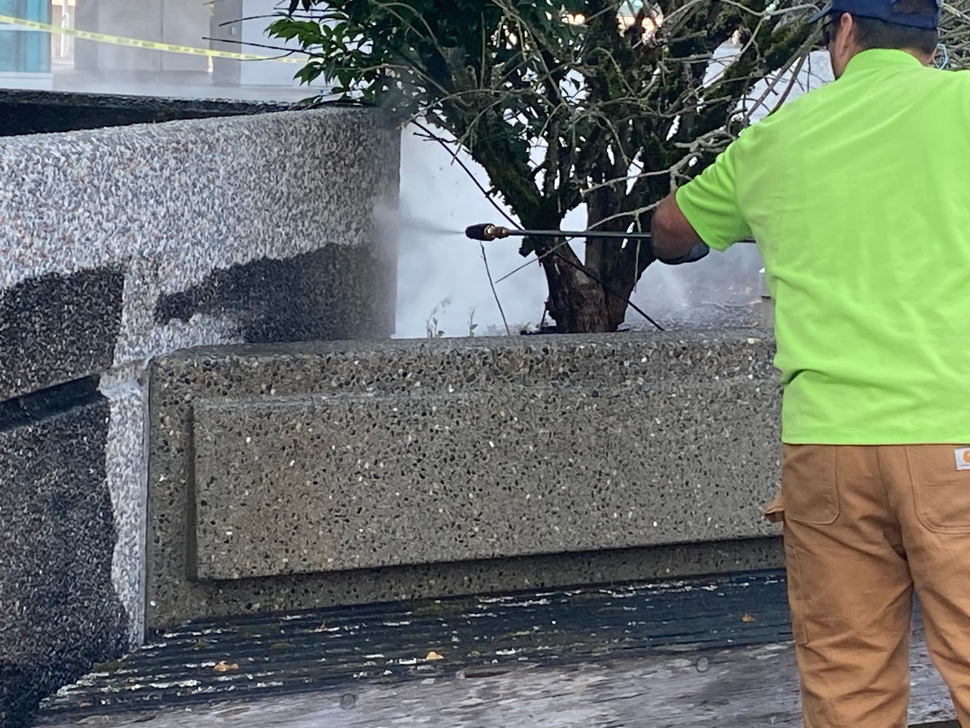 Worker in neon green shirt spraying water onto a concrete planter near a tree.