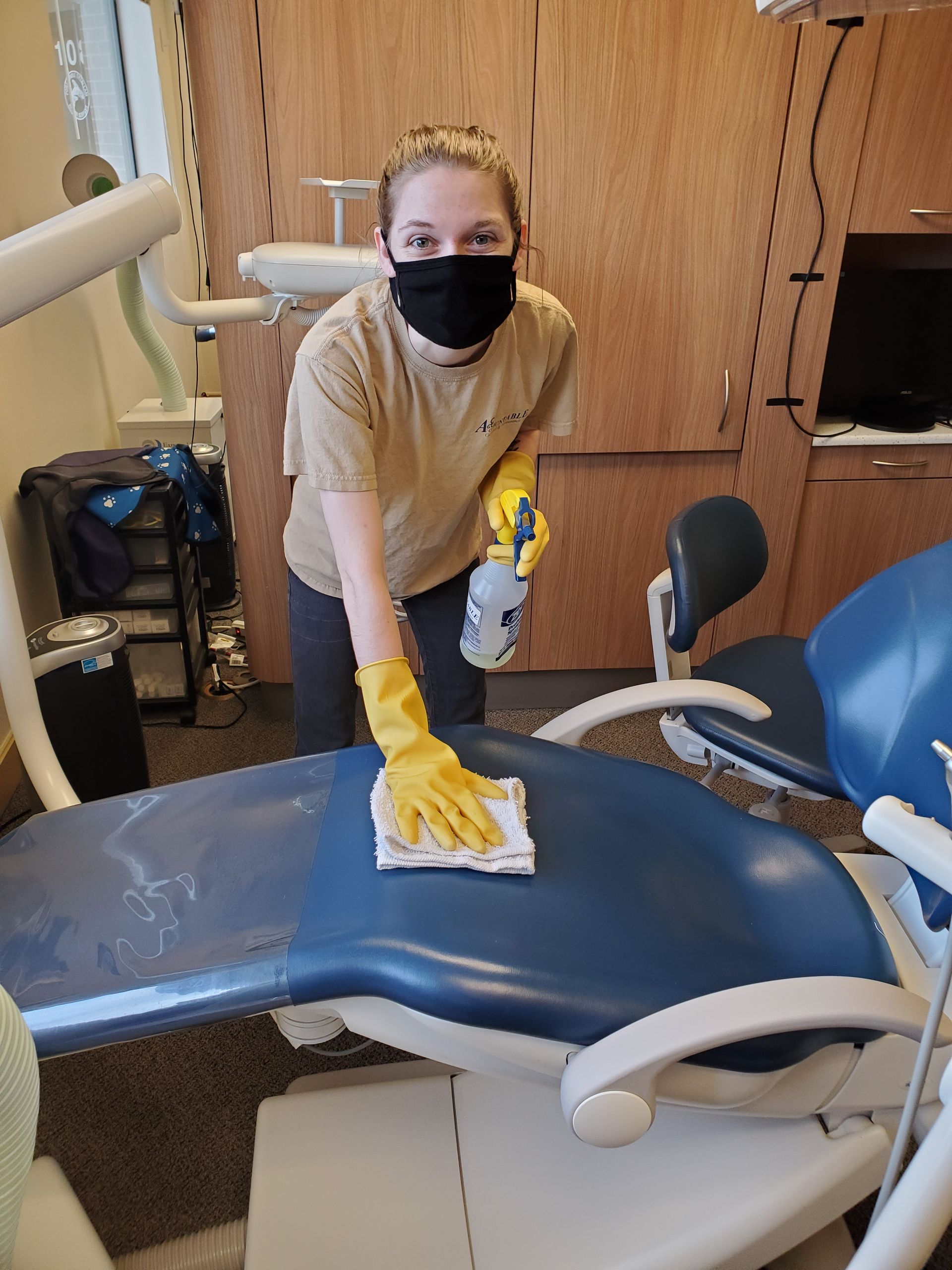 Masked worker wipes a dental chair with a cloth in a clinic room.