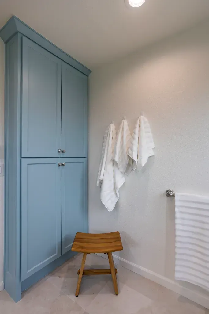 A bathroom with blue cabinets , a wooden stool , and towels hanging on the wall.