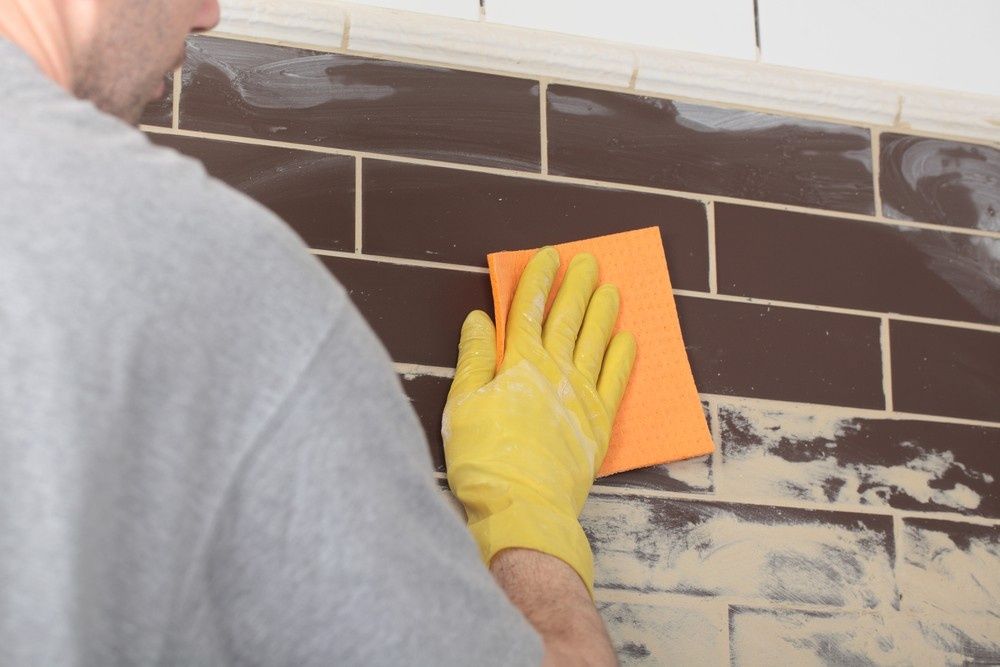 Person Wearing a Yellow Glove is Wiping Grout Off Dark Brown Tiles With an Orange Sponge — North Coast Tiles in Grafton, NSW