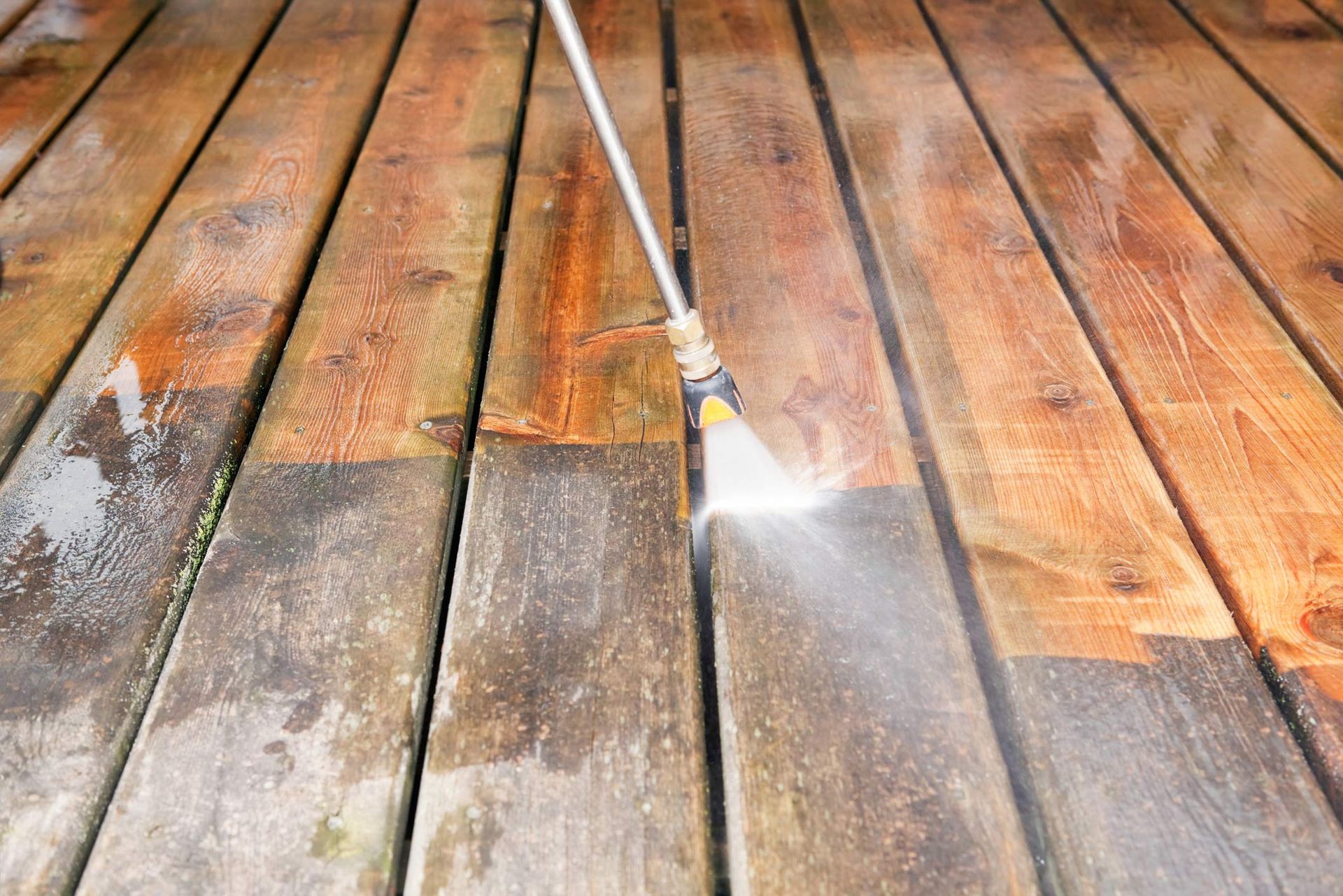 Power washer cleaning a weathered wooden deck, revealing a brighter, cleaner surface.