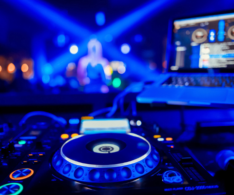 Blue-lit DJ turntable and laptop on a stage, with blurred performer and bright concert lights in the background