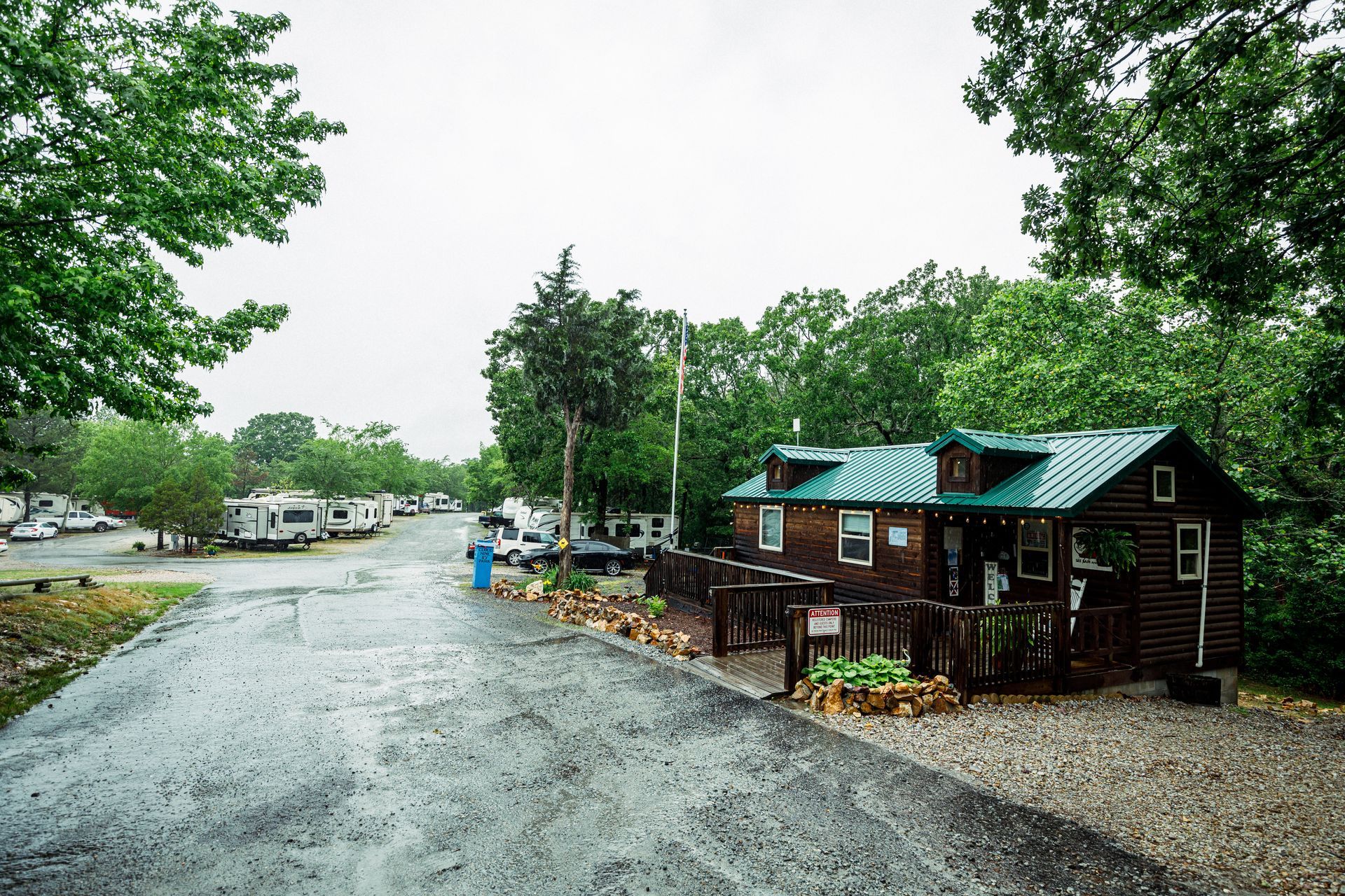 Rainy campground with a gravel road, trees, parked vehicles, and small cabins.