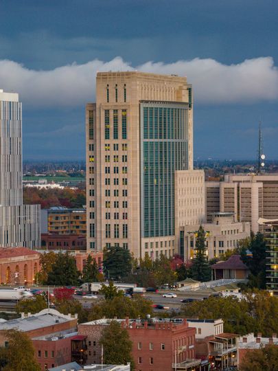 Tall beige building with glass facade, surrounded by shorter buildings and autumn trees, under a cloudy sky.