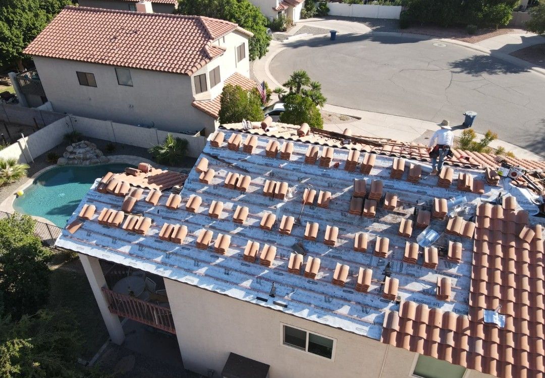Roofer on a house roof replacing red clay tiles. Other roofed houses and a swimming pool are visible.